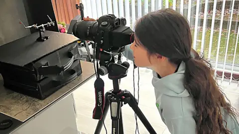 Family photo A girl with long brown tied-back hair, wearing a light blue hoodie and looking through a black camera on a tripod.