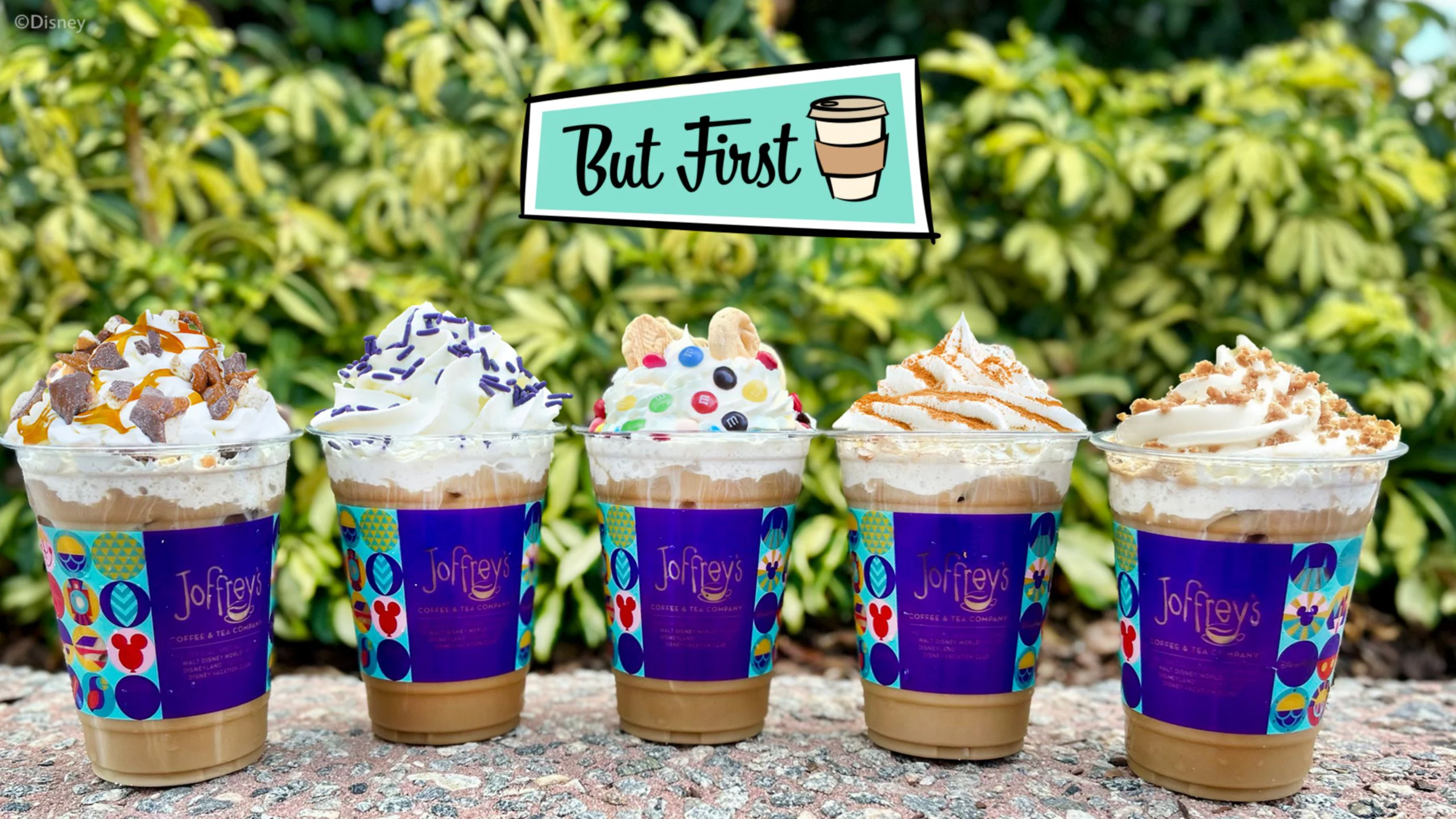 Five iced coffees with whipped cream and toppings in colorful cups, with a sign above reading “But First” and a coffee cup.