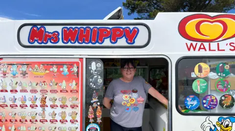 BBC Sarah McKeich stood inside an ice cream van. She is smiling at the camera, and is wearing a striped top with badges on it. There are pictures of ice creams on the windows.