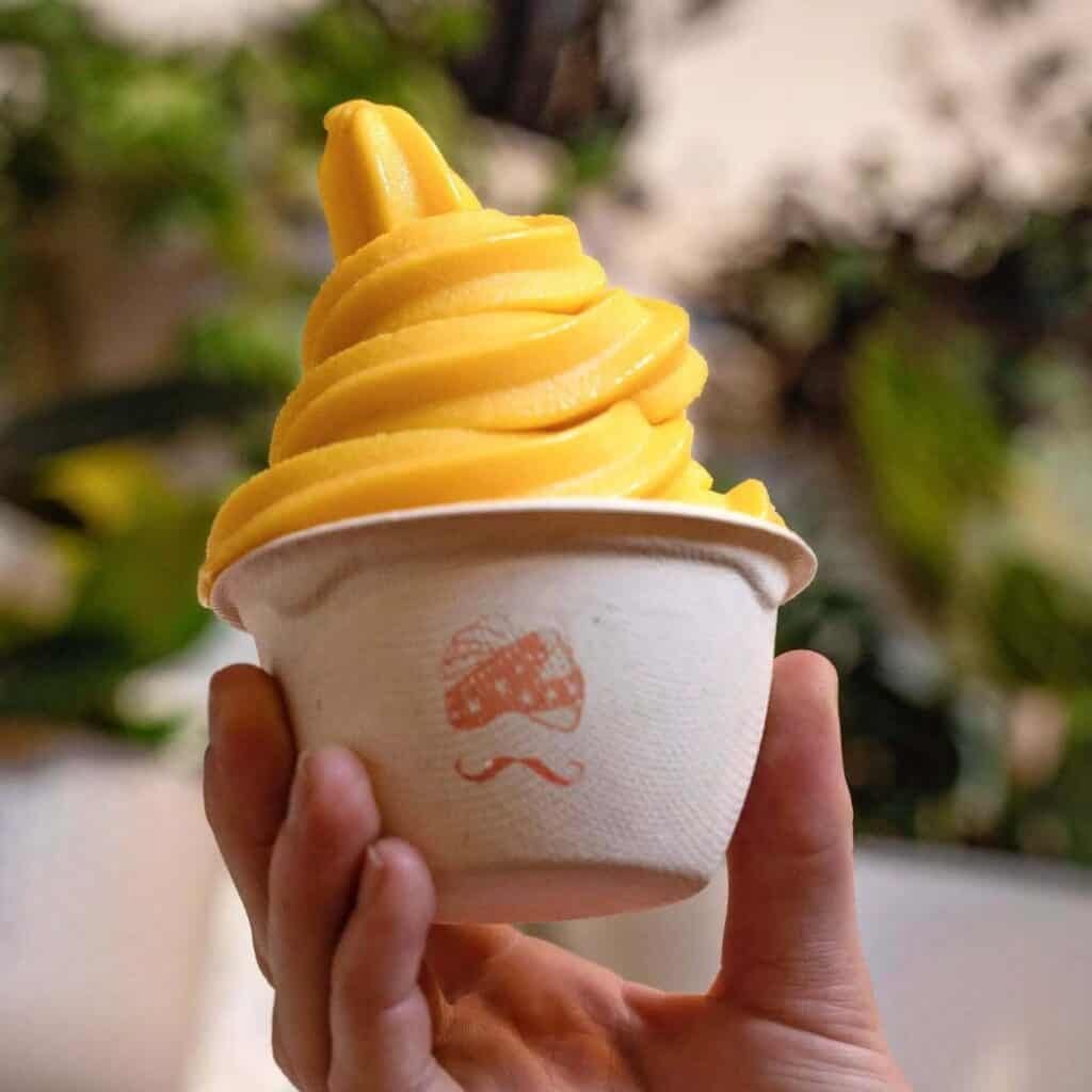 A hand holds a cup of yellow soft-serve ice cream against a blurred background with green foliage.