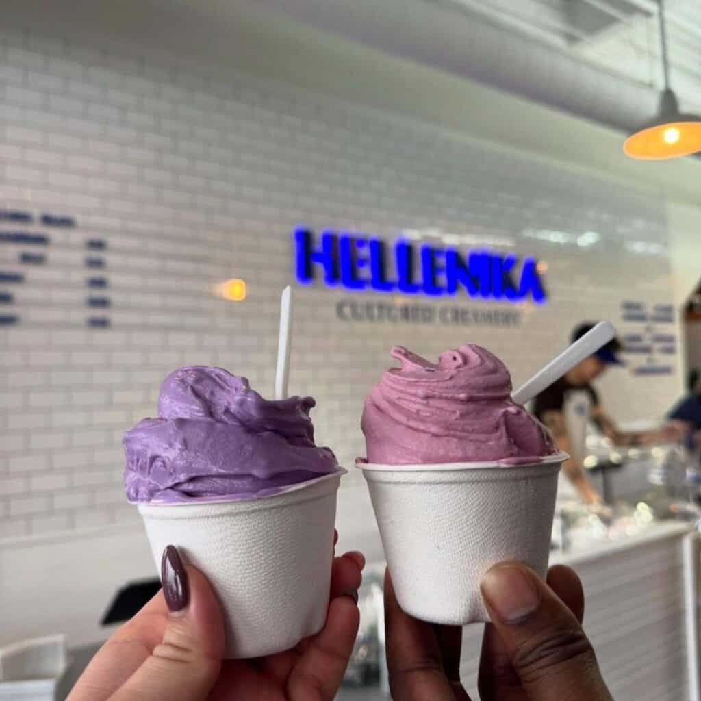 Two hands holding cups of purple and pink gelato in front of a white tiled wall with a blue "HELLENIKA" sign in the background.