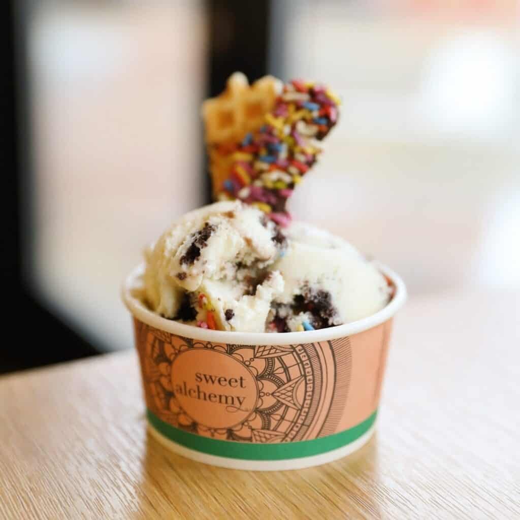 A cup of cookies and cream ice cream topped with a waffle piece coated in chocolate and sprinkles, on a wooden table.