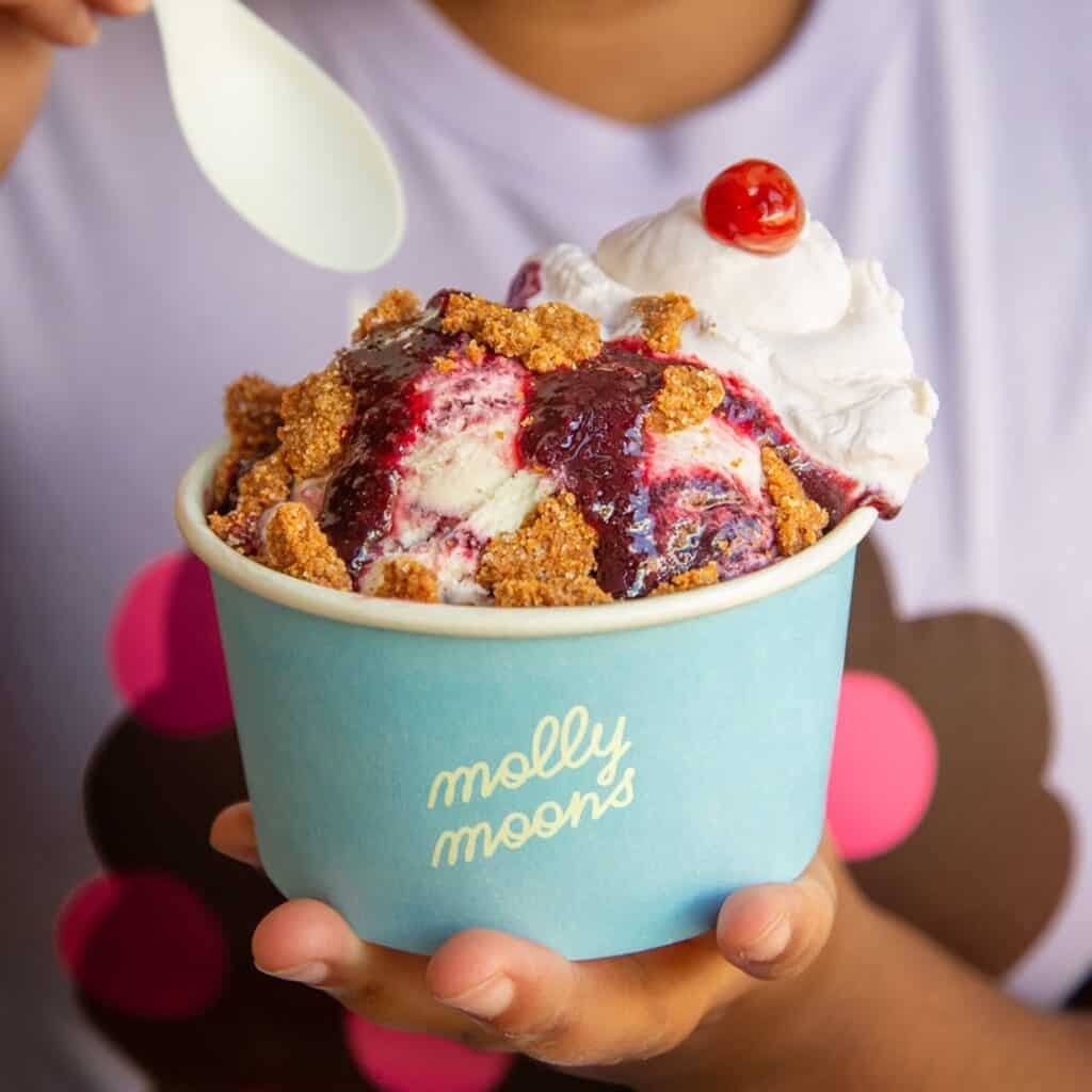 A person holds a cup of ice cream topped with berry sauce, whipped cream, crumbled cookie, and a cherry. The cup reads "molly moons.