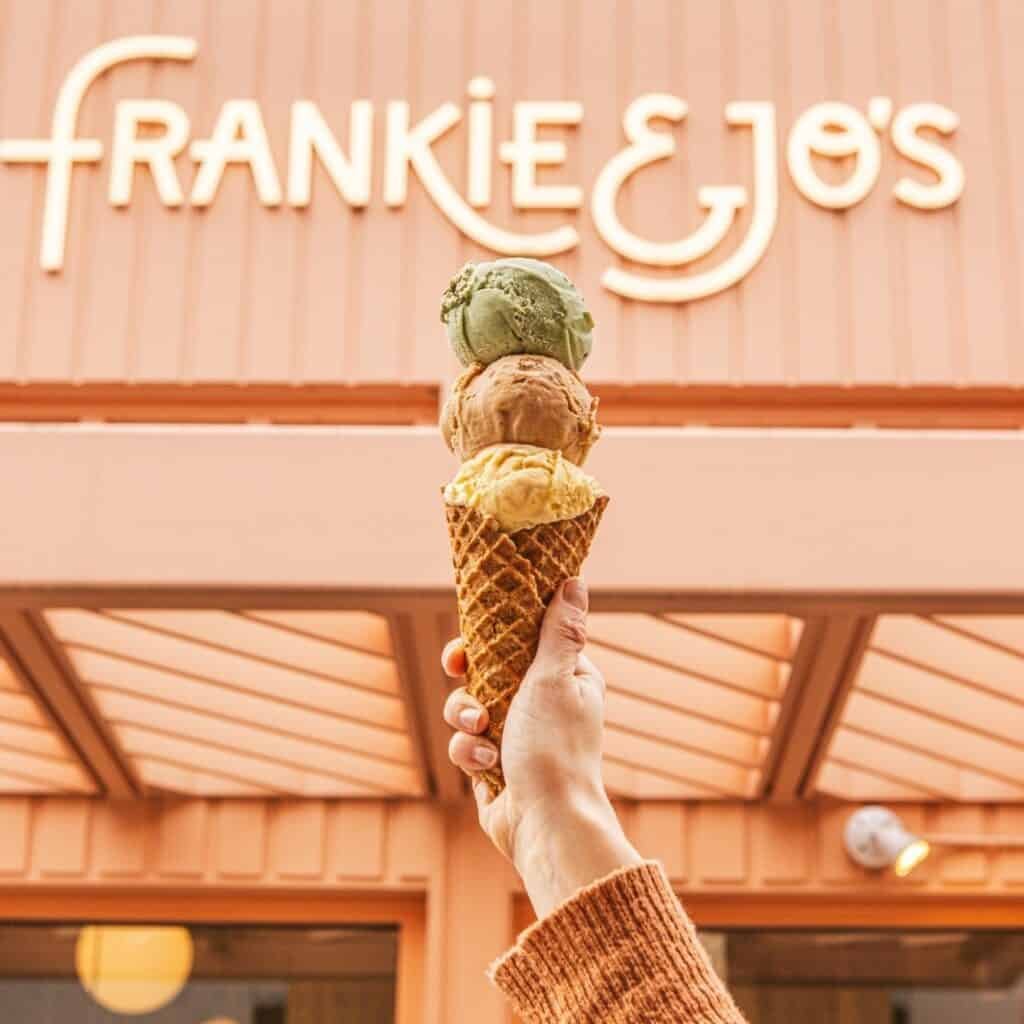 A hand holds a triple-scoop ice cream cone in front of a peach-colored storefront with the sign "FRANKIE & JO'S.