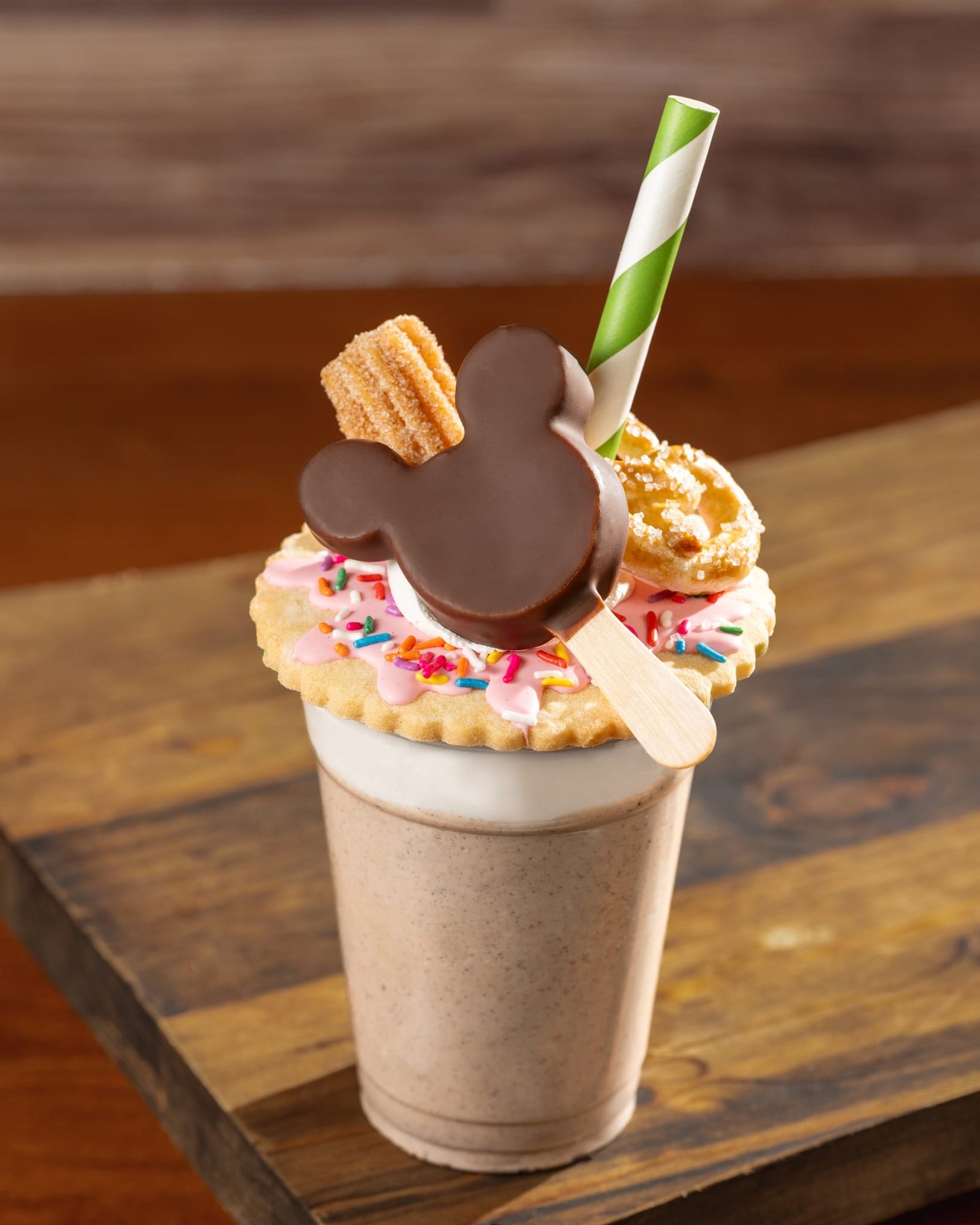 A milkshake topped with a cookie, pink frosting, sprinkles, churros, a Mickey-shaped chocolate, and a green striped straw.